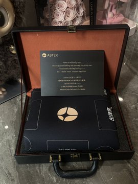 A black presentation box with the 'ASTER' logo contains a card and a dark, tech-themed item, with the visible text reading 'Thank you for being part of our story since the very beginning. We hope you enjoy your journey with ASTER. THE MORE THE ASTER, THE BRIGHTER WE SHINE.'