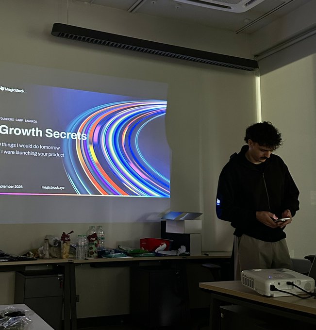 A young man in a black hoodie standing in a dimly lit room looking at his phone, with a projection screen behind him displaying a MagicBlock presentation titled 'Growth Secrets' from a Bangkok founders camp.