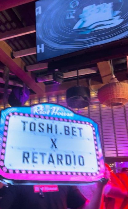 A photo taken inside a venue showing a neon-lit marquee sign that reads "Red House" at the top with "TOSHI.BET X RETARDIO" displayed below, with screens and lighting fixtures visible in the background.