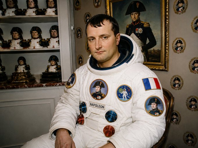 A portrait of a man in a white astronaut suit labeled 'NAPOLEON', decorated with patches depicting Napoleon Bonaparte, set against a background filled with Napoleon-themed busts and portrait plates.