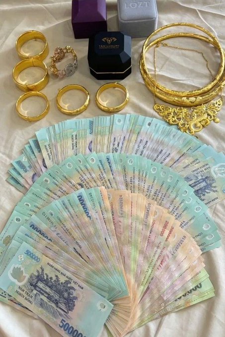 A display of gold jewelry including bangles, rings, and necklaces, along with jewelry boxes labeled 'TAMA LOYALTY', arranged above a large fan of Vietnamese Dong banknotes with denominations of 500,000 VND visible.