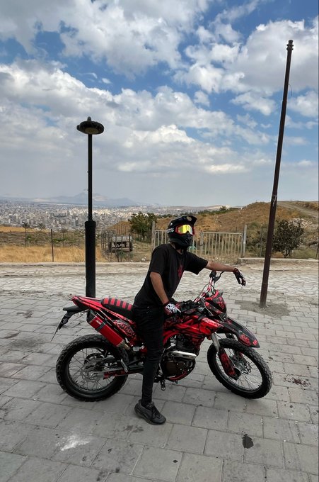 A person wearing a black helmet and outfit sitting on a red and black dirt bike on a paved overlook with a city view in the background.
