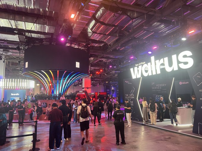 A crowded trade show floor featuring a large illuminated 'wallrus' booth displaying 'MEMORY SERVERS' and a colorful light installation with rainbow LED streams.