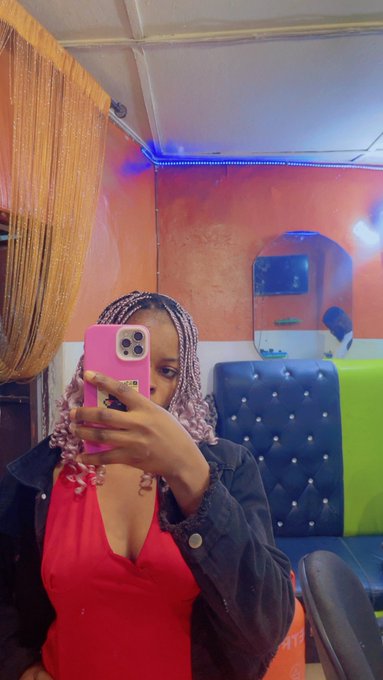 A woman with purple braided hair takes a mirror selfie with a pink phone while wearing a red top and denim jacket in a lounge with blue seating and orange walls.