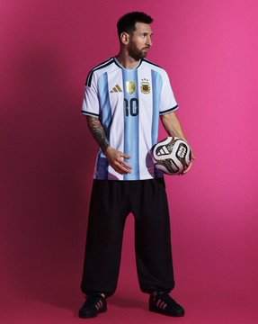 A man wearing an Argentina national football team jersey with the number 10 (Messi's number) holding a soccer ball, standing against a solid pink/magenta background.
