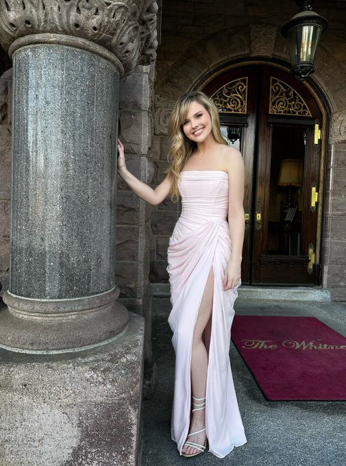 A young blonde woman in a pink strapless gown with a high slit stands at the entrance of 'The Whitley' building, leaning against a stone column.