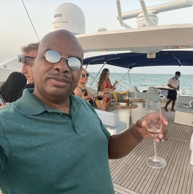 A man in a teal polo shirt and aviator sunglasses smiles while holding a glass of champagne on the deck of a luxurious yacht, which features a "SeaVision" satellite dome and "$80k" worth of upscale surroundings including plush seating and ocean views.