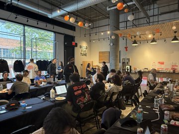 A group of around 30 attendees sits at long tables inside a bright, modern event space, working on laptops during the @bankrbot hackathon. White paper lanterns hang from the ceiling, and a partially visible sign in the background reads "BUILD ELLA TUNE," reflecting the balanced-gender turnout the post highlights.