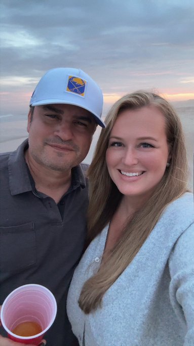 A selfie of a smiling couple on a beach at sunset; the man wears a light blue cap with a mountain patch and a gray shirt, while the woman has long blonde hair and wears a gray top. A pink plastic cup is visible in the foreground.
