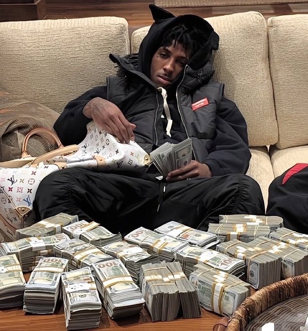 A young man wearing a black puffer jacket with the hood up sits on a couch, casually holding and counting stacks of U.S. dollar bills while surrounded by numerous additional bundles of cash.