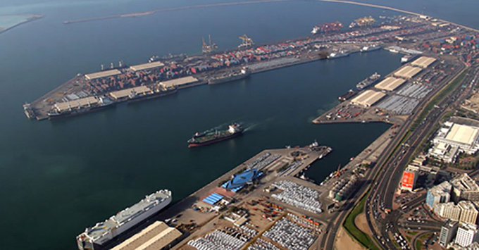 An aerial photograph of a large industrial seaport with multiple cargo ships docked at extensive container terminals, shipping yards filled with stacked containers, and a cityscape visible in the background.