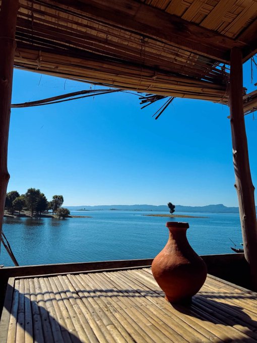 A scenic view from under a bamboo shelter, looking out over a calm blue lake toward small tree-covered islands, with a terracotta pot resting on a wooden deck.
