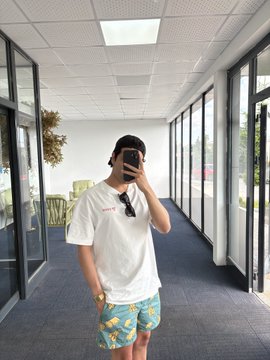 A person in a white t-shirt with a red logo and blue floral shorts takes a mirror selfie in a bright office hallway, showcasing their casual summer outfit. The red logo on the shirt aligns with the post's reference to a "familiar red logo," fitting the warm-weather vibe of the #TronGlobalFriends #TGF theme.