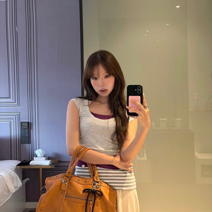 A mirror selfie of a young woman with long brown hair wearing a white tank top, holding a pink phone case and an orange handbag in what appears to be a hotel room.