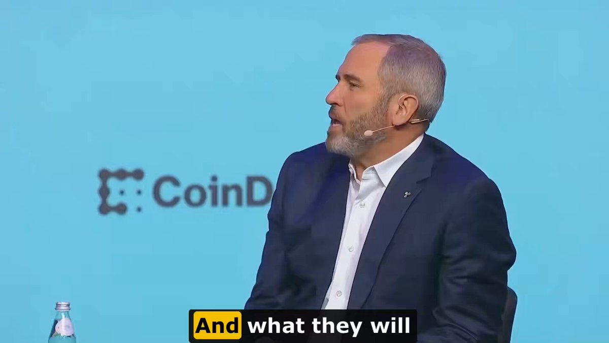 The image shows a man wearing a suit, sitting at a table with his hands on his lap while talking to someone. The background features a blue wall with the word "CoinD" written on it. Below this, there are two lines of text that read "And what they will".