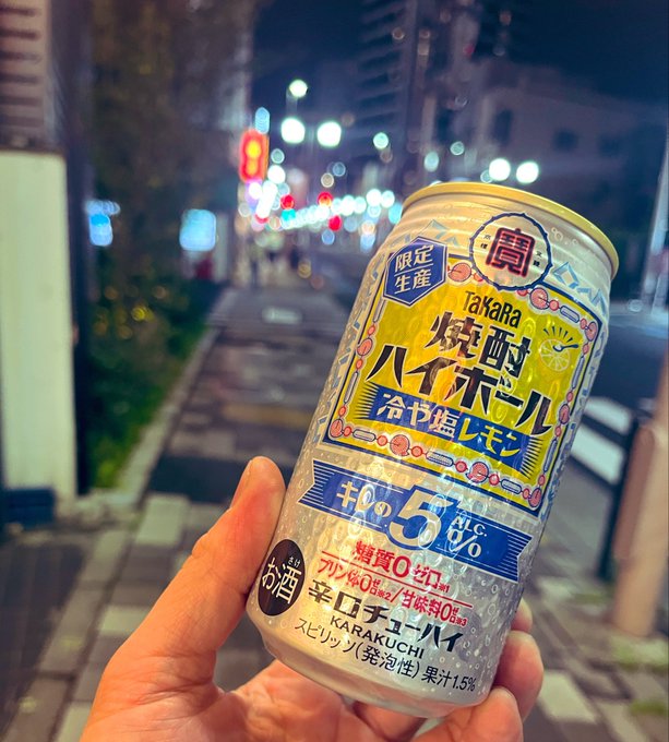 The image features a hand holding up a can of Takara beer, which has Japanese writing on it. The background shows a city street at night with lights illuminating the scene. The photo appears to be taken from a low angle, making the beer can appear larger and more prominent in the frame.