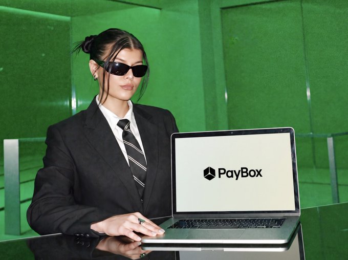 A woman in a black suit and sunglasses sits at a desk using a laptop that displays the 'PayBox' logo, set against a green paneled backdrop.