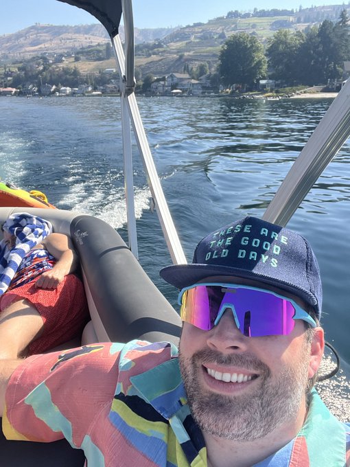 A man wearing reflective sunglasses and a hat reading 'THESE ARE THE GOOD OLD DAYS' takes a selfie while boating on a lake, with hills visible in the background.