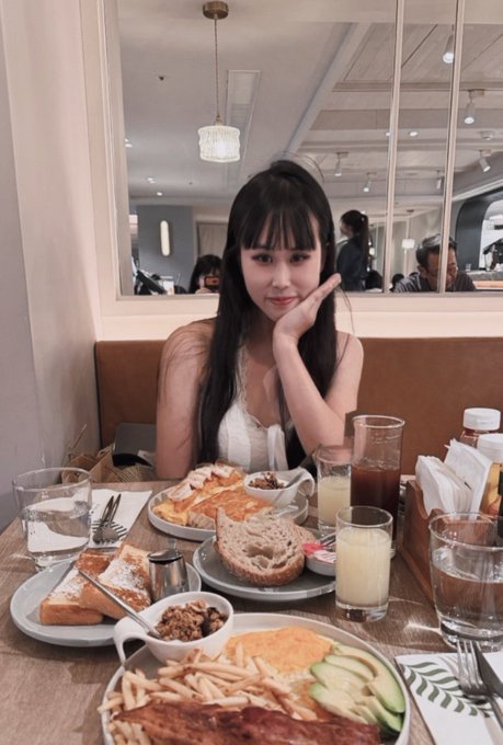 A young woman with long dark hair poses at a restaurant table filled with various dishes including bread, fries, avocado, and meats, with drinks and condiments also visible.