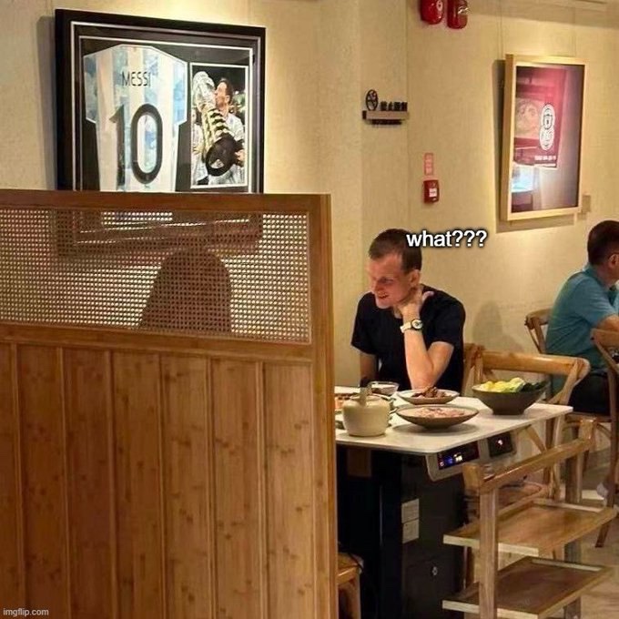 A man eating at a restaurant looks up in surprise at a framed Lionel Messi #10 jersey on the wall, with the caption 'what???' edited above his head.