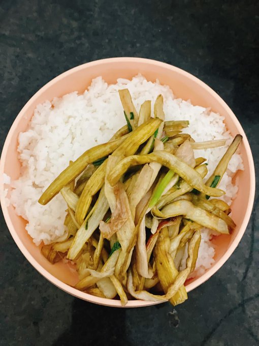 Overhead view of a peach-colored bowl filled with white rice topped with stir-fried vegetables, likely oyster mushrooms or eggplant, served on a dark countertop.