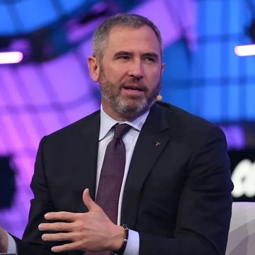 Brad Garlinghouse, CEO of Ripple, is shown speaking at what appears to be a conference or panel event, wearing a dark suit with a maroon tie, with his hand gesturing mid-sentence. The image accompanies news of his critical comments about Strategy's (formerly MicroStrategy) financial engineering practices negatively impacting the crypto market.