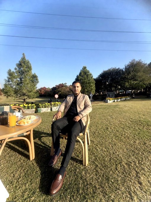 A well-dressed man in a beige jacket, black shirt, dark pants, and brown loafers sits on a wooden chair beside a small table with juice and snacks on a sunny grassy lawn, perfectly capturing a relaxed yet polished "good morning, ready to make big wins" vibe.