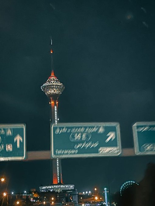 A nighttime photograph featuring an illuminated telecommunications tower, resembling the Milad Tower, with overhead street signs displaying directional arrows in both English and Persian/Farsi script.