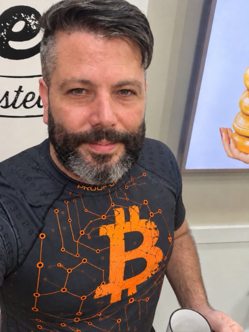 A bearded man wearing a black Bitcoin "Proof of Work" t-shirt takes a selfie, embodying the disciplined daily routine of waking early, stacking sats, training jiu-jitsu, and praising the Lord. The prominent orange Bitcoin logo on his shirt visually reinforces his commitment to dollar-cost averaging into BTC as part of his morning ritual.