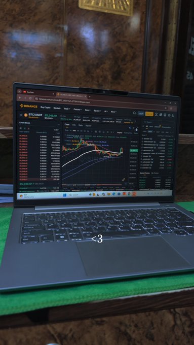 A gray laptop sits on a wooden table displaying a Binance trading chart for the BTC/USDT pair on its screen.
