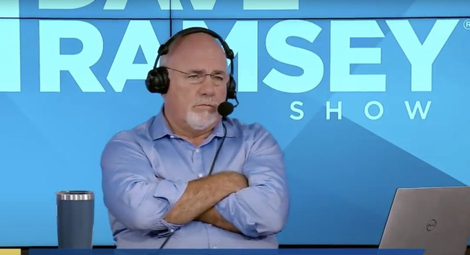 A bald man with a white beard wearing glasses and a blue shirt sits with arms crossed wearing headphones in front of a "RAMSEY SHOW" backdrop.