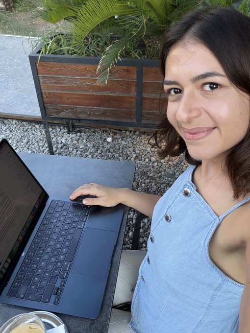 A woman with dark shoulder-length hair wearing a light blue denim top smiles while working on a dark blue laptop at an outdoor table, with a wooden planter box visible behind her.