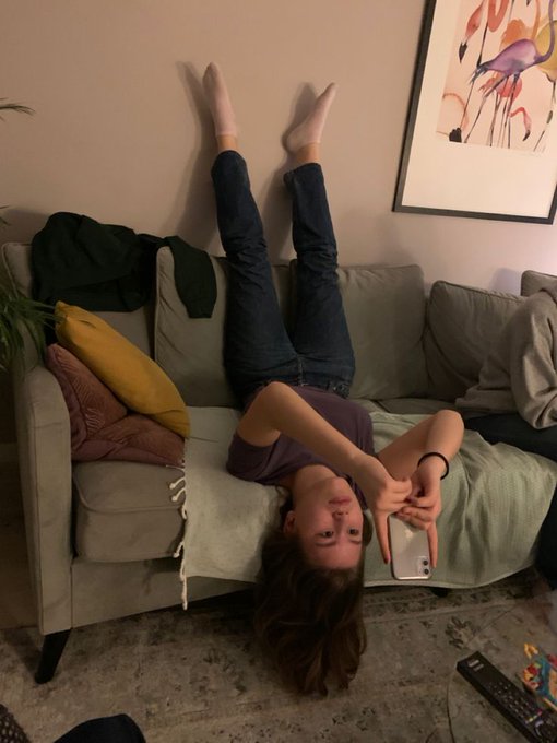 A young woman lying upside down on a couch with her legs up against the wall, holding a smartphone and smiling at the camera.