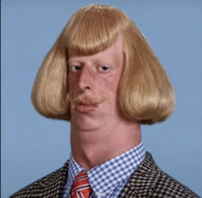 A person with fair skin, freckles, and a blonde bowl-cut hairstyle with a curled mustache poses against a blue background, wearing a blue and white checkered shirt, red striped tie, and tweed blazer.
