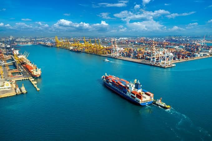 An aerial view of a large container ship being assisted by a tugboat at a busy port with numerous yellow cranes loading and unloading shipping containers.