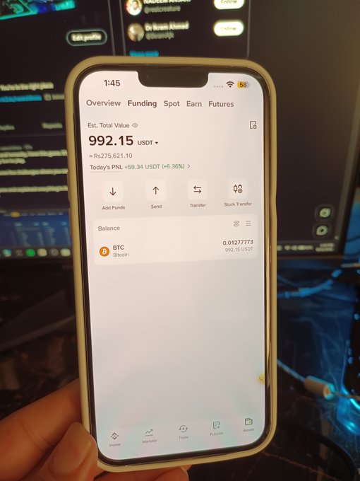 A hand holding a smartphone displaying a cryptocurrency exchange app (appears to be Bybit) showing an estimated total value of 992.15 USDT, with a BTC balance of 0.01277773 BTC, and a today's PnL of +59.34 USDT (+6.36%).