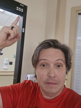 The image features a man wearing a red shirt, with his face turned towards the camera as he points at something outside of the frame. The background includes a mirror and a door, suggesting that this scene takes place within an indoor space like a bathroom or bedroom. The caption reads "Let' s riiiiippppp!!! After 16 days in the ICU room @ 555 (no effing joke!!), finally home and ready to rip some @veefriends !!! Thanks for the love and support @garyvee @jeremyknowsVF @VeeFriendsCards".
