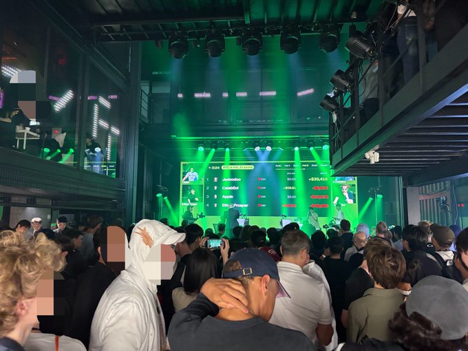 A crowded indoor event with green stage lighting, where a large screen displays a leaderboard with names like 'Jimboboo' and 'GreenSkull' along with crypto values.