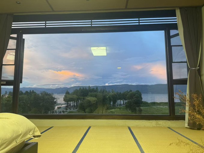 The image shows a tranquil view through a large window of a lake surrounded by mountains at sunset, with tatami mat flooring and traditional Japanese-style interior framing the scenic vista. The serene landscape image appears unrelated to the accompanying promotional text about creator incentives on OKX Planet.