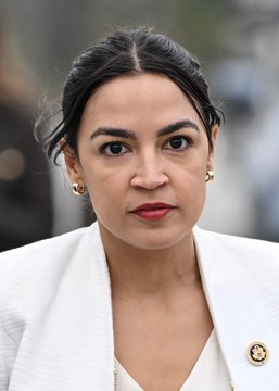 A close-up portrait of a woman with dark hair pulled back, wearing a white blazer with a decorative brooch and red lipstick, looking toward the camera.