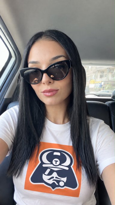 A selfie of a woman with long black hair wearing cat-eye sunglasses and a white t-shirt with an orange graphic design, taken inside a car.