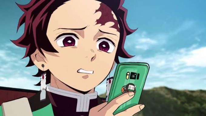 An anime character (Tanjiro from Demon Slayer) with a concerned expression holding a green phone displaying a 'Genshin Impact' character on the screen.