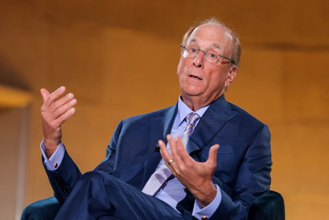 A photograph of a bald man with glasses wearing a blue suit and patterned tie, sitting in a chair and gesturing with his hands as if speaking during a presentation.