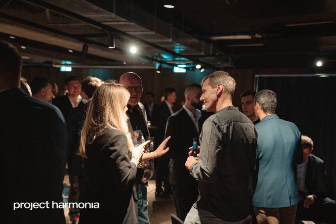 A photograph of an indoor networking event with people in business attire conversing under dim lighting, labeled 'project harmonia' in the bottom left corner.