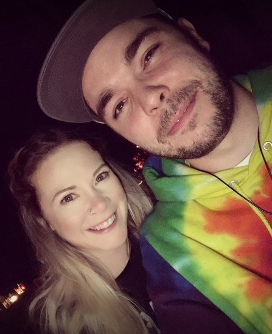 A young couple poses for a nighttime selfie, with the man wearing a colorful tie-dye hoodie and a baseball cap, while the woman with long blonde hair smiles beside him. The image's casual, fun vibe aligns with the post's playful "degen" persona embracing the crypto culture, though no specific bitcoin-related text or tickers are visible in the photo itself.