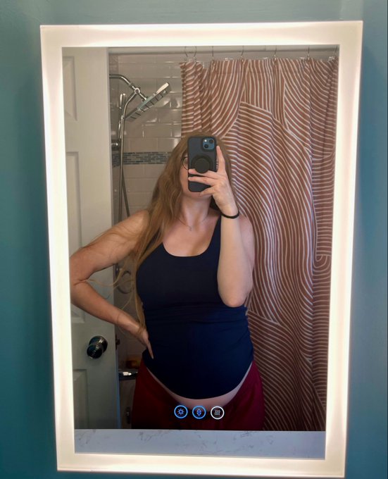 A mirror selfie shows a woman in a navy tank top and maroon pants, with a visible baby bump that aligns with the post's "Happy bump day btw" caption celebrating her pregnancy. The mirror also displays camera app icons at the bottom for adjusting photo settings.