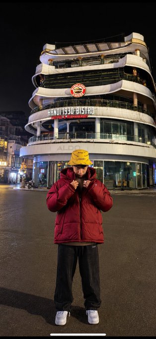 A person in a red puffer jacket and yellow bucket hat stands in front of an illuminated multi-story building displaying 'Hanoi 1930 Bistro' and 'Aldo' signage at night.