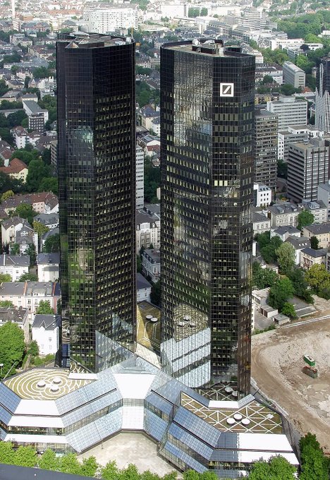 An aerial view shows two tall dark twin skyscrapers (one with a Deutsche Bank logo) rising above a glass-roofed structure and surrounding cityscape.