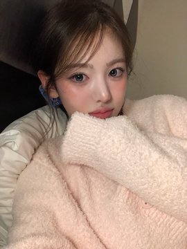 A young woman wraps herself in a plush cream-colored knit sweater or blanket, with a visible blue earbud in her ear as she gazes softly at the camera. The image relates to the post "260930 #Sui Bubble," suggesting the cozy, bubbly texture of her sweater embodies the warm and soft aesthetic associated with the Sui Bubble trend.