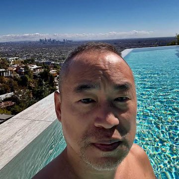 A close-up selfie of a shirtless man with short hair, smiling slightly, overlooking a cityscape with a large blue swimming pool in the background.